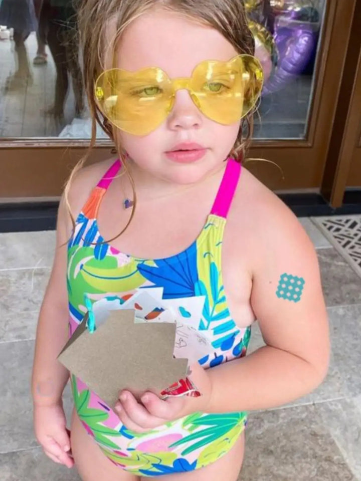 A young girl wearing yellow heart-shaped sunglasses shows an applied NATPAT Magic Patch: Natural Bug Bite, Itch Relief Stickers on her arm | Twentyseven Toronto