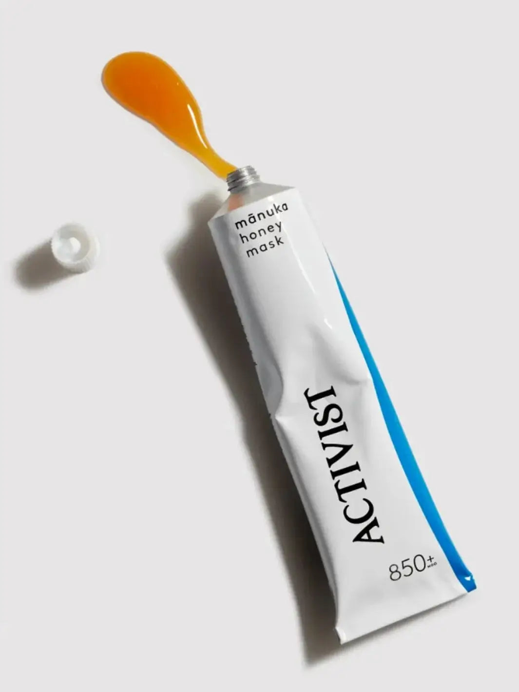 A white squeeze tube of Activist Manuka Mānuka Honey Mask lies flat against a plain grey surface with a dollop of golden honey product oozing from the top next to its detached cap | Twentyseven Toronto
