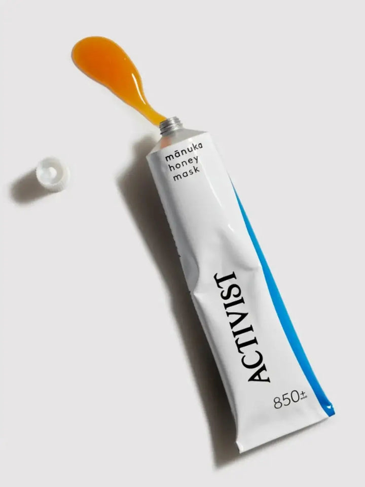 A white squeeze tube of Activist Manuka Mānuka Honey Mask lies flat against a plain grey surface with a dollop of golden honey product oozing from the top next to its detached cap | Twentyseven Toronto