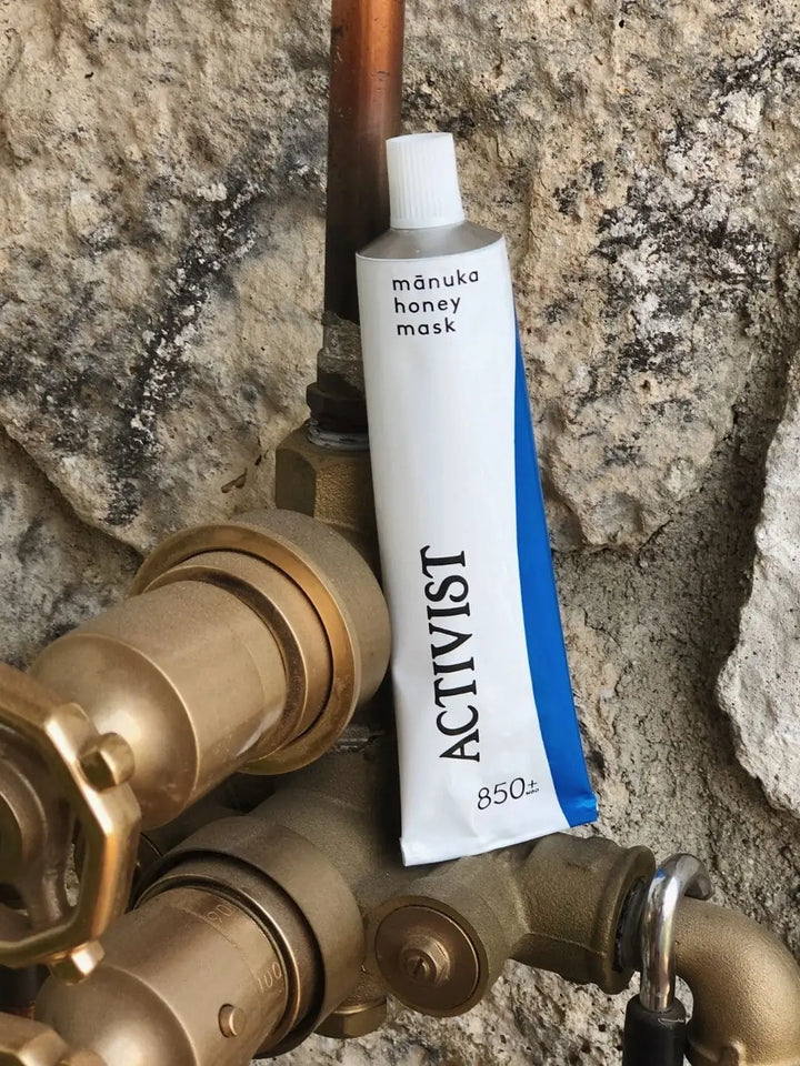A tube of Activist Manuka Mānuka Honey Mask stands upright, wedged behind rustic, industrial brass pipes against a textured stone wall | Twentyseven Toronto