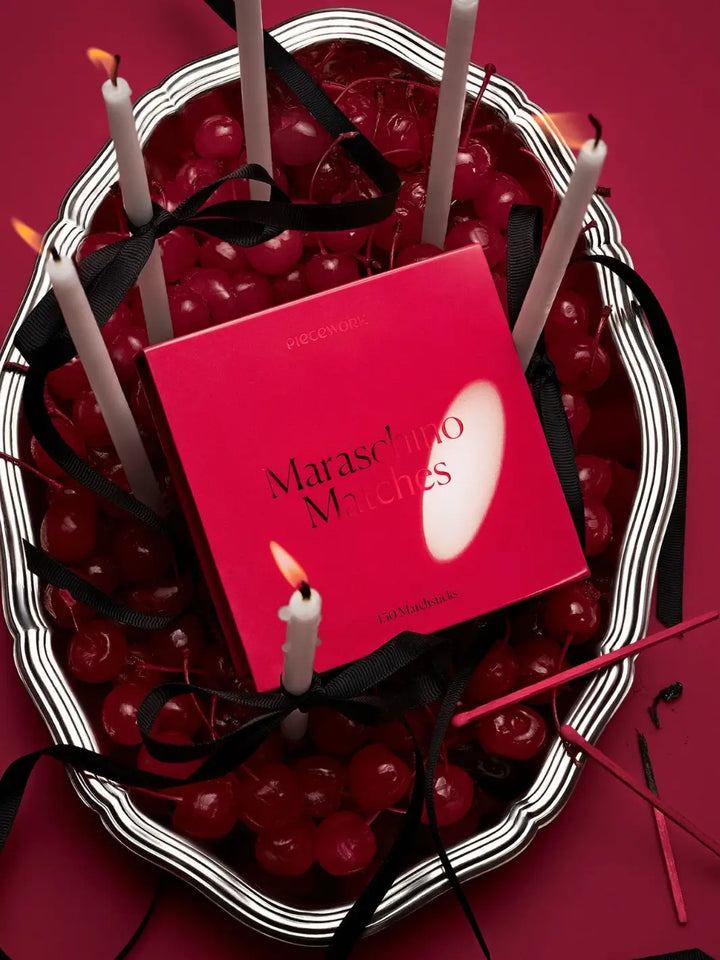 Silver basket full of maraschino cherries with lit candles and a red box labeled 'Maraschino Matches' from Piecework Puzzles on a red background | Twentyseven Toronto