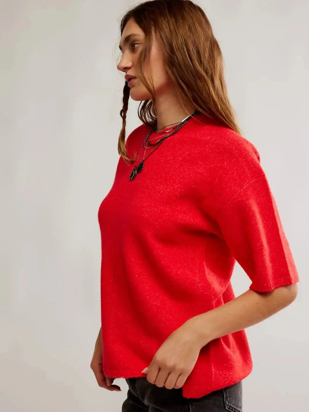 A front view shows a model posing with hands on her hips, wearing the vibrant Free People Margot SoftSpun Sweater Tee in Cherry Pop Melange accessorized with layered silver necklaces and black jeans | Twentyseven Toronto