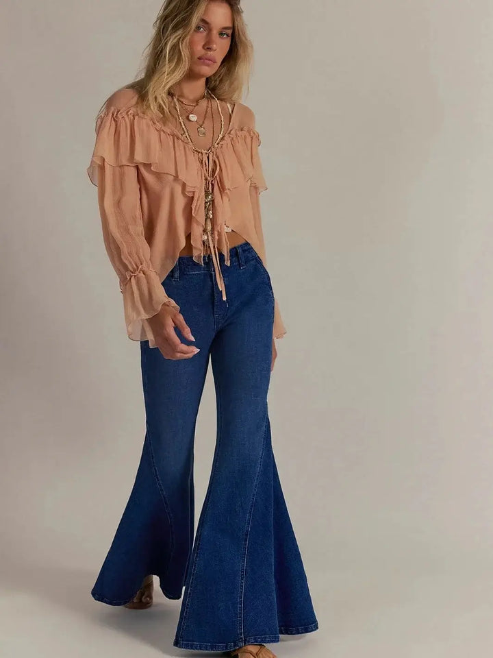 A model poses from the front wearing the Free People Marque Flare Jeans - Kora styled with a ruffled, sheer long-sleeve blouse and layered necklaces | Twentyseven Toronto
