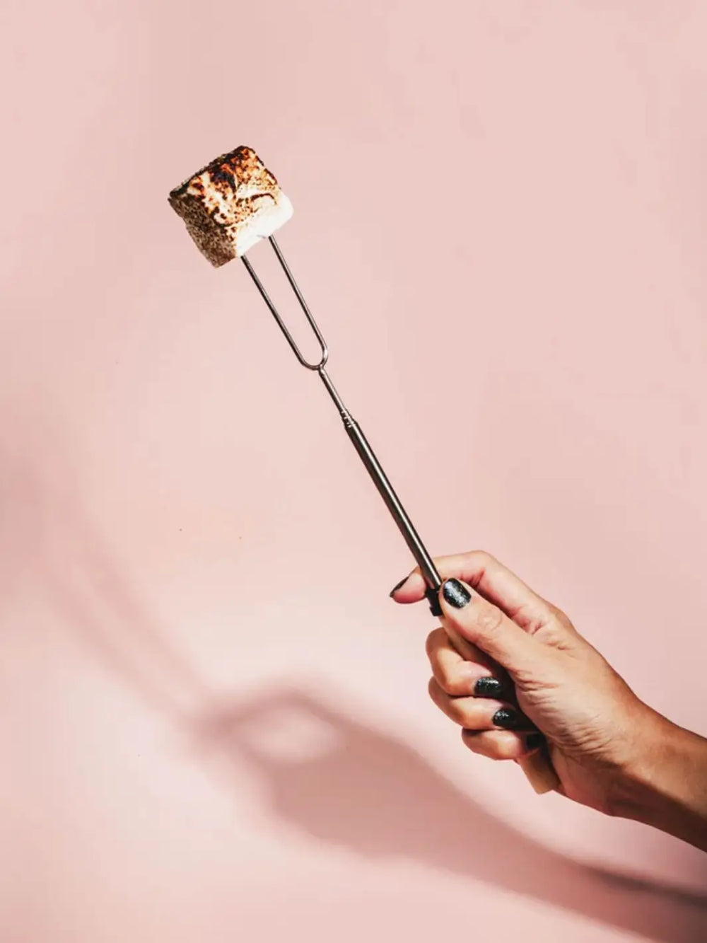 A hand with dark sparkly nail polish holds a Mojave Mallows Marshmallow Skewer / Roasting Stick with a perfectly toasted, golden-brown marshmallow on its prongs against a plain pink background | Twentyseven Toronto