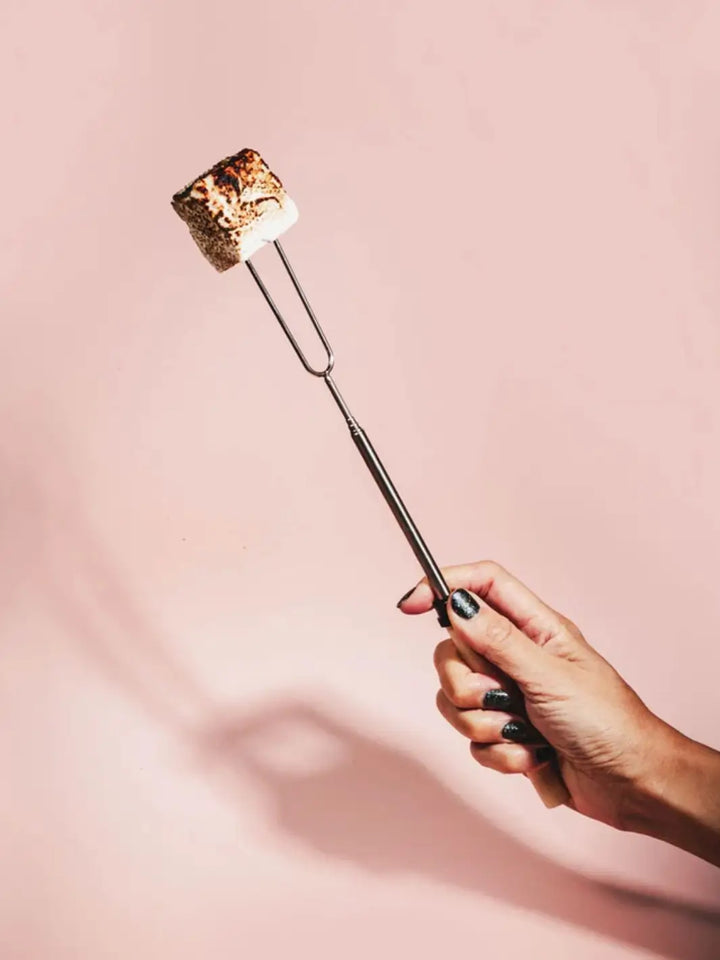 A hand with dark sparkly nail polish holds a Mojave Mallows Marshmallow Skewer / Roasting Stick with a perfectly toasted, golden-brown marshmallow on its prongs against a plain pink background | Twentyseven Toronto