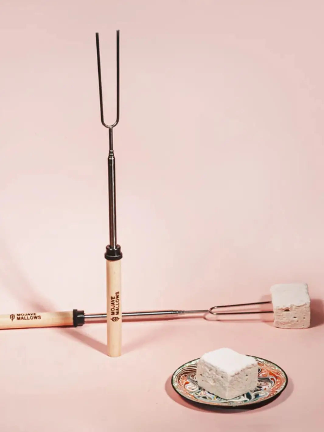 A vertical Mojave Mallows Marshmallow Skewer / Roasting Stick stands upright next to another one lying flat with a square marshmallow attached, placed near a small decorative plate on a soft pink surface | Twentyseven Toronto