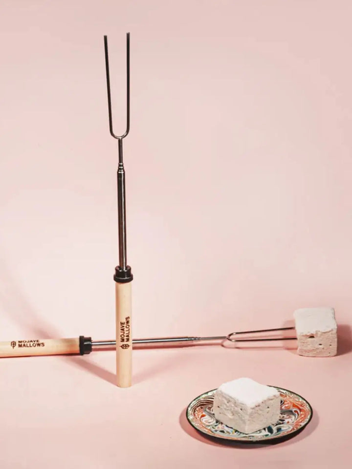 A vertical Mojave Mallows Marshmallow Skewer / Roasting Stick stands upright next to another one lying flat with a square marshmallow attached, placed near a small decorative plate on a soft pink surface | Twentyseven Toronto