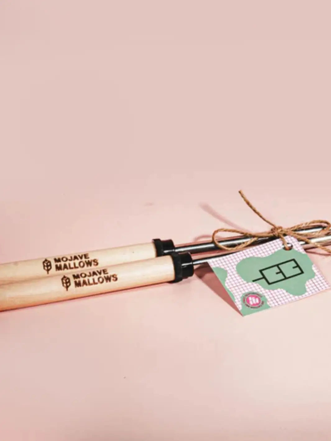 Two metal telescoping tools with branded wooden handles are tied together with twine alongside a small checkered product tag, showcasing the Mojave Mallows Marshmallow Skewer / Roasting Stick set against a pink backdrop | Twentyseven Toronto