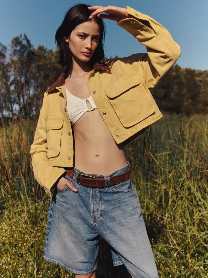An outdoor shot shows a model wearing the unbuttoned Free People Martha Workwear Applique Jacket in Rattan over a white bikini top and slouchy denim shorts in a grassy field | Twentyseven Toronto