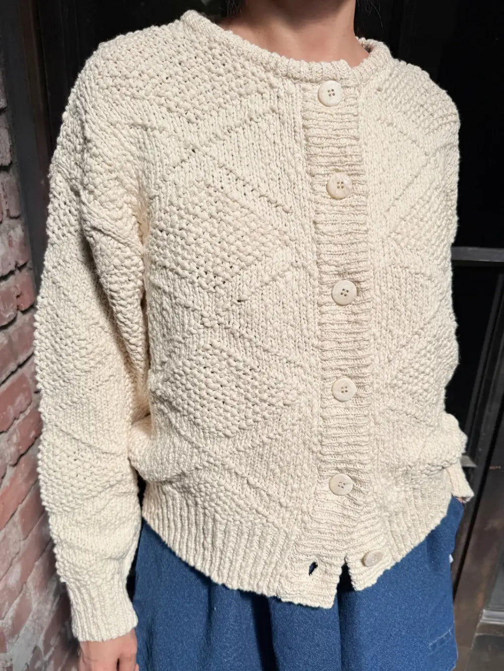 Person wearing a beige knitted 'Mason Cotton Cardigan' in 'Naturel' with buttons standing against a dark background | Twentyseven Toronto