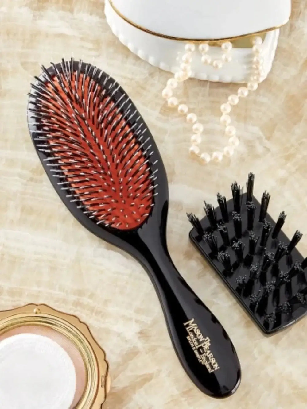 A premium black hairbrush with white nylon bristles resting on a red rubber pad is displayed on a vanity table alongside a pearl necklace and a small black Mason Pearson Cleaning Brush CL | Twentyseven Toronto