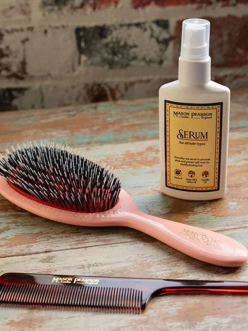 A white pump bottle of Mason Pearson Hair Serum stands on a rustic wooden table alongside a classic pink hairbrush and a dark tortoiseshell comb | Twentyseven Toronto
