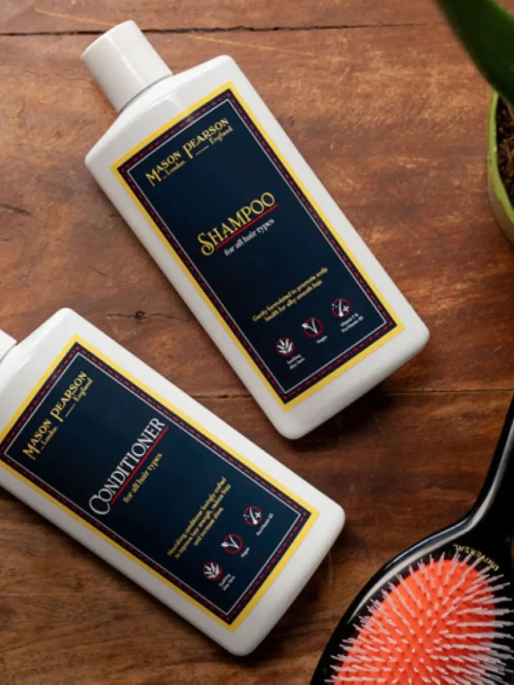 A white bottle of shampoo and a matching white bottle of conditioner are placed on a warm wooden surface next to a classic black hairbrush to display the premium Mason Pearson Shampoo and Conditioner collection | Twentyseven Toronto