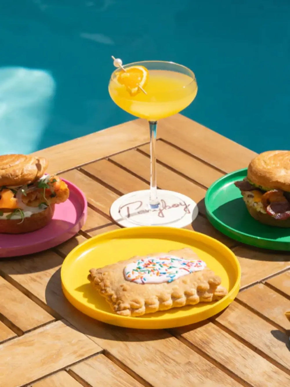 Three plates from the Heller Max Dinnerware 9.75 inch Maxplate in Rainbow - Set of 6 hold breakfast food and sandwiches on a wooden table next to a pool | Twentyseven Toronto