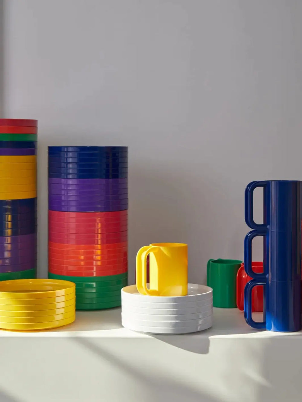 A collection of colourful plates, bowls, and stacking mugs from the Heller Max Dinnerware Set in Rainbow, arranged neatly on a white shelf | Twentyseven Toronto