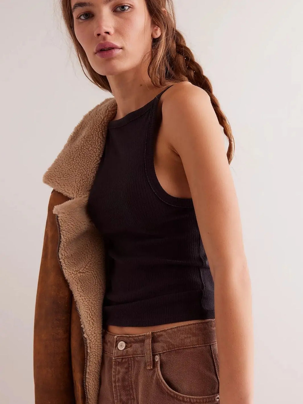 A model with braided hair showcases the high square neckline and slim fit of the Free People Mia Tank - Black styled with a brown shearling jacket resting on her shoulder | Twentyseven Toronto