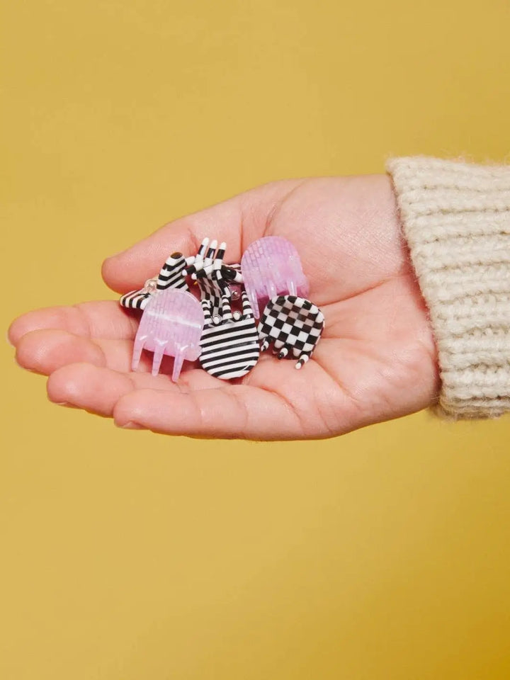 A hand holds a loose pile of the six tiny checkered, lilac, and striped CHUNKS Micro Claws in Geo against a mustard yellow background | Twentyseven Toronto
