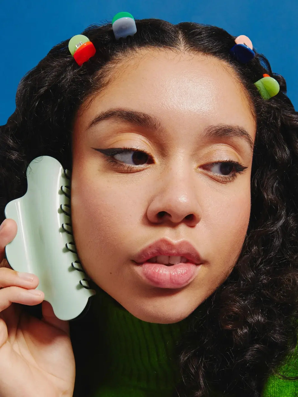 A close-up of a person with curly dark hair wearing several colorful hair accessories, including the Chunks Hairclips Micro Claws in Play set pinned along their hairline while they hold a larger mint green claw clip next to their face | Twentyseven Toronto