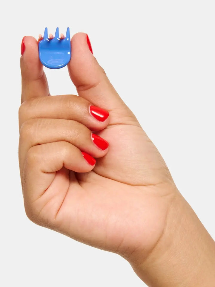 A hand with bright red fingernails holds up a single tiny, blue claw clip from the Chunks Hairclips Micro Claws in Play collection against a solid light gray background | Twentyseven Toronto
