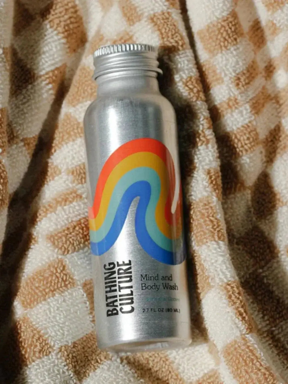 A small aluminum bottle of Bathing Culture Mind & Body Wash - Cathedral Grove (Travel Size) features a colorful rainbow graphic and rests on a brown and white checkered towel | Twentyseven Toronto