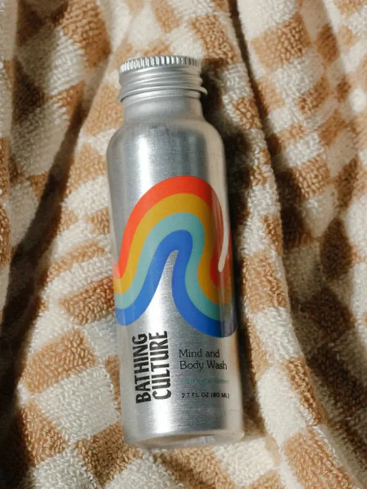 A small aluminum bottle of Bathing Culture Mind & Body Wash - Cathedral Grove (Travel Size) features a colorful rainbow graphic and rests on a brown and white checkered towel | Twentyseven Toronto