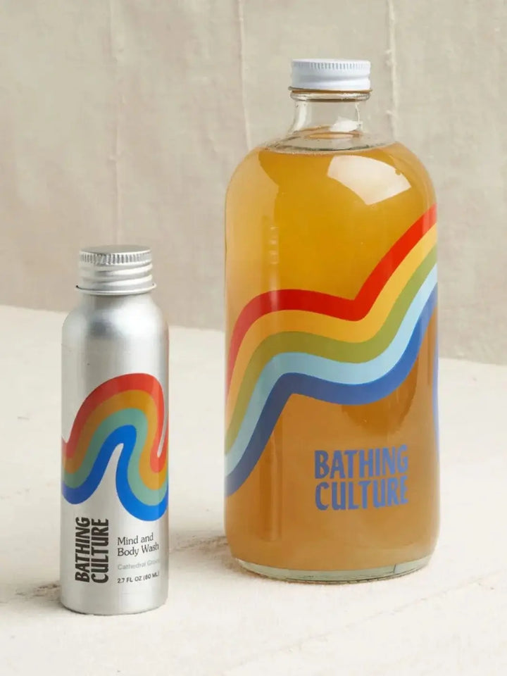 A side-by-side comparison displays a small aluminum bottle of Bathing Culture Mind & Body Wash - Cathedral Grove (Travel Size) next to a large glass refill bottle filled with amber liquid gel | Twentyseven Toronto