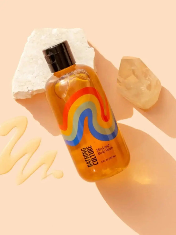 An amber bottle of Bathing Culture Mind & Body Wash - Meadow Vision rests on a peach background alongside a streak of liquid gel and decorative natural stones | Twentyseven Toronto