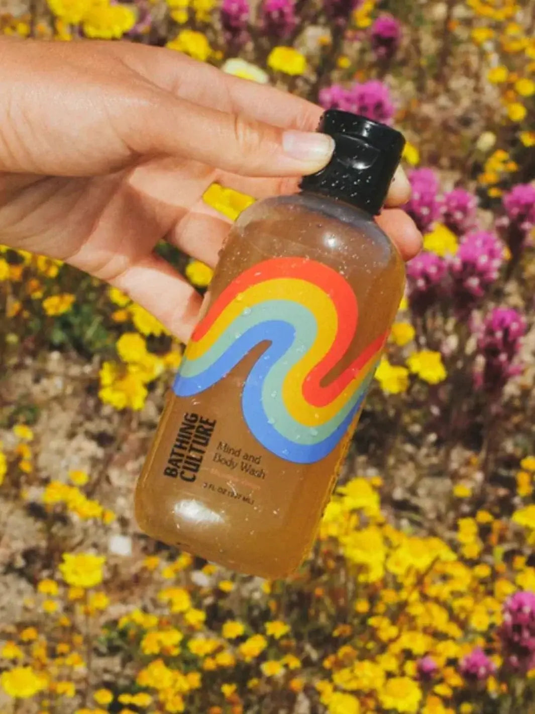 A hand holds an amber travel bottle of Bathing Culture Mind & Body Wash - Meadow Vision against a vibrant backdrop of yellow and purple wildflowers in a field | Twentyseven Toronto