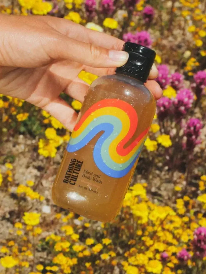 A hand holds an amber travel bottle of Bathing Culture Mind & Body Wash - Meadow Vision against a vibrant backdrop of yellow and purple wildflowers in a field | Twentyseven Toronto