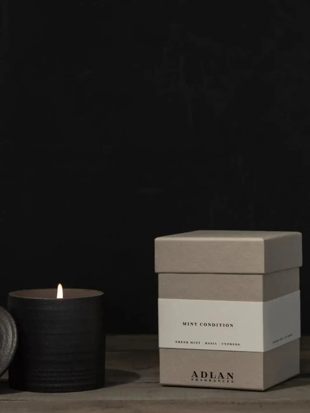 A lit Adlan Fragrances Mint Condition - Petite candle glows inside its textured black jar next to its grey product box against a dark backdrop | Twentyseven Toronto