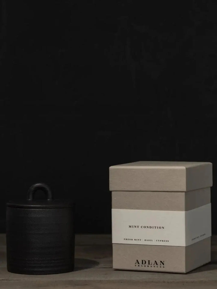 The Adlan Fragrances Mint Condition - Petite candle sits covered with its matching black lid next to its square grey packaging box on a wooden table | Twentyseven Toronto