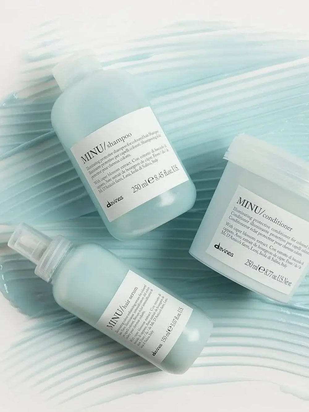 A bottle of Davines MINU Hair Serum lies diagonally alongside a matching shampoo and conditioner tub, all arranged over a textured background of light blue product smears | Twentyseven Toronto