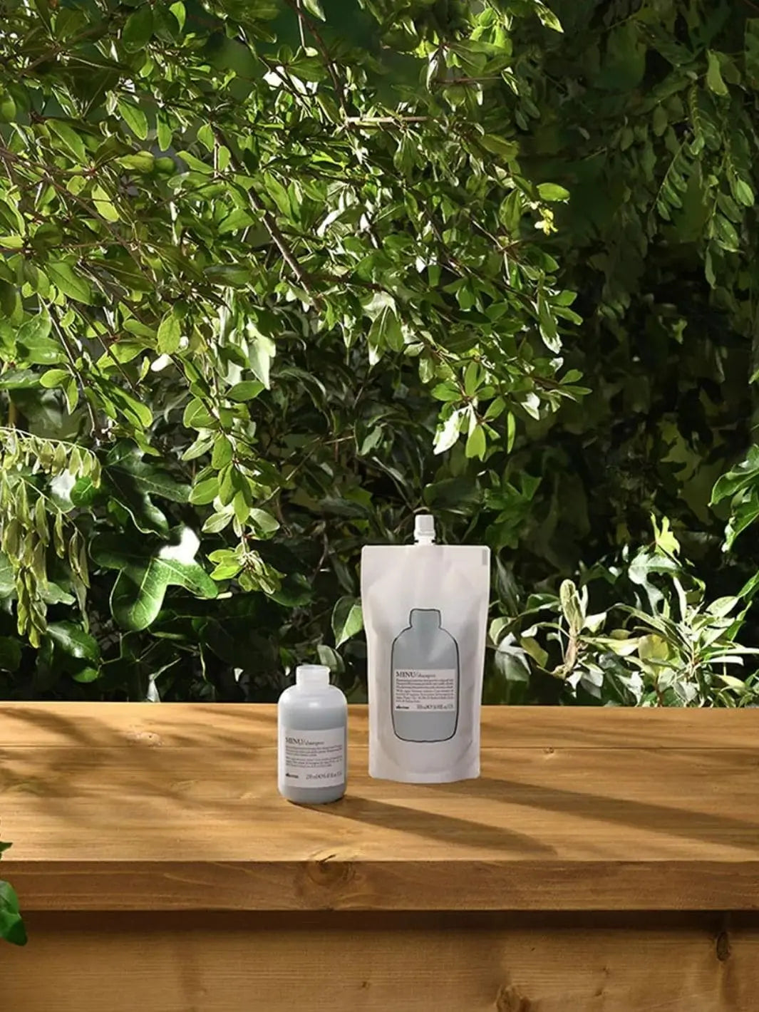 A pouch of Davines Shampoo Refill and a small plastic bottle rest on a wooden table with lush green foliage blurred in the background | Twentyseven Toronto