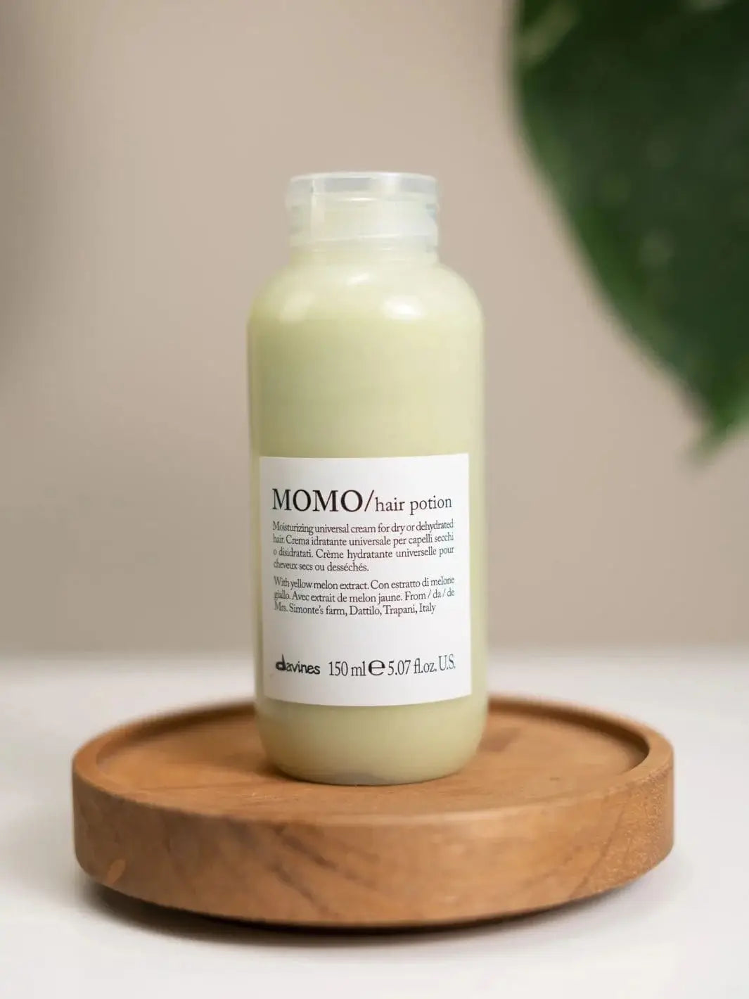 A bottle of Davines MOMO Hair Potion stands on a small, circular wooden tray against a neutral background | Twentyseven Toronto