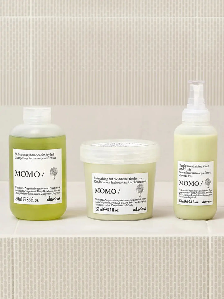 A product group shot shows the Davines Momo Serum spray bottle sitting on a light tiled ledge alongside its matching moisturizing shampoo and fast conditioner for dry hair | Twentyseven Toronto