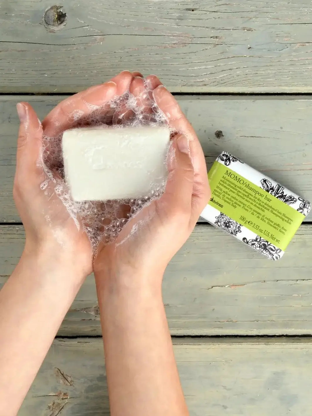 A pair of hands covered in white soap suds holds an unwrapped solid bar next to a paper-packaged Davines MOMO Shampoo Bar on a rustic wooden surface | Twentyseven Toronto