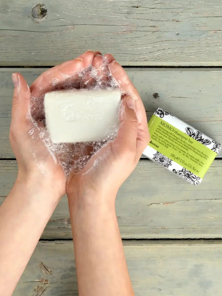 A pair of hands covered in white soap suds holds an unwrapped solid bar next to a paper-packaged Davines MOMO Shampoo Bar on a rustic wooden surface | Twentyseven Toronto