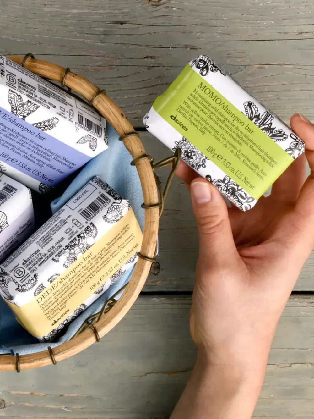 A hand lifts a green-labeled Davines MOMO Shampoo Bar out of a small woven basket filled with other wrapped shampoo bars | Twentyseven Toronto