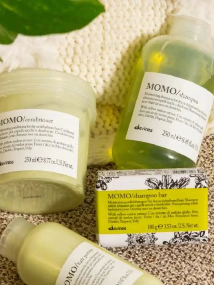 Several bars of paper-wrapped Davines MOMO Shampoo Bar sit stacked together on a dark cloth inside a woven basket | Twentyseven Toronto