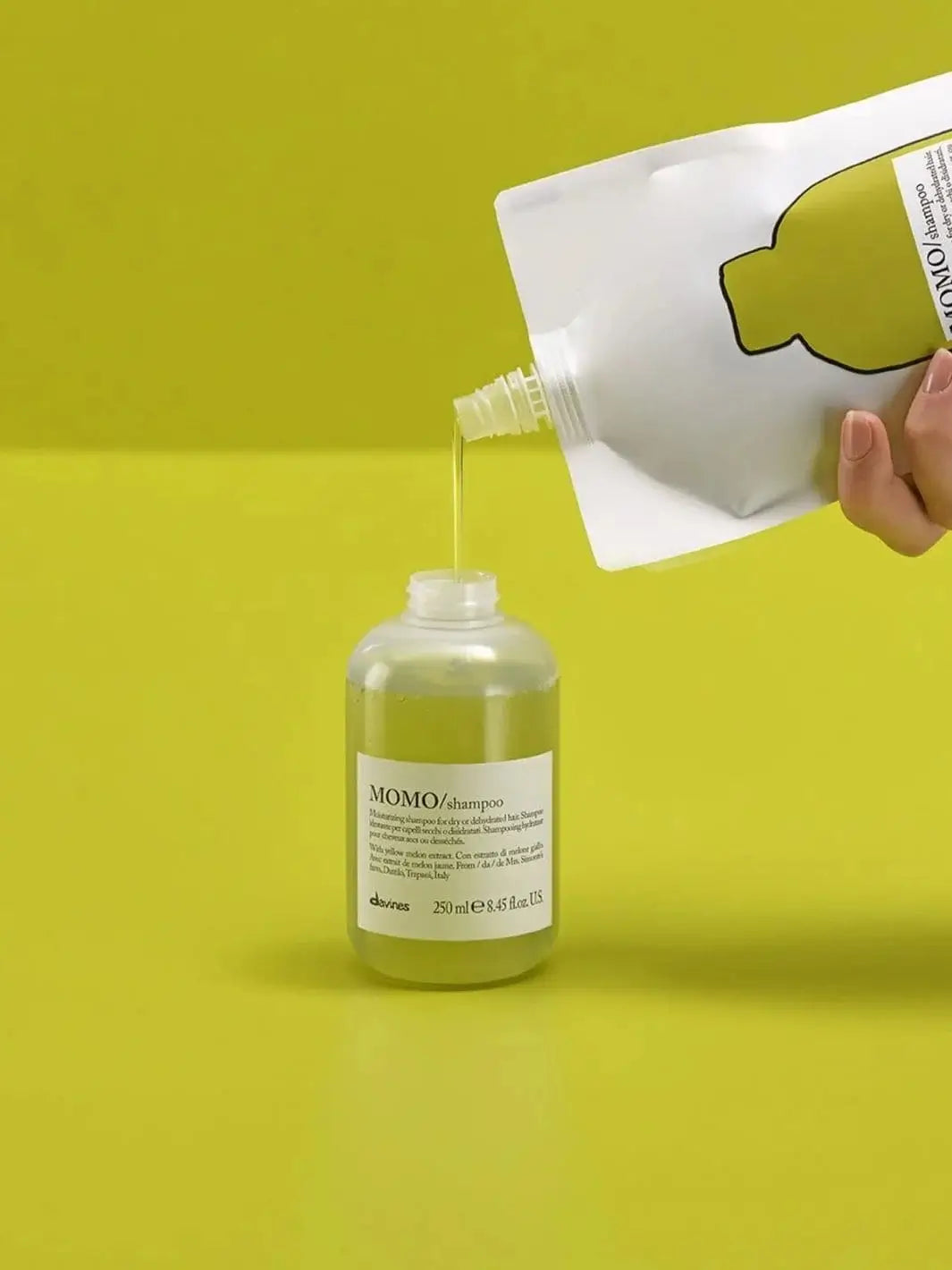 A hand pours translucent green liquid from a white pouch of Davines MOMO Shampoo Refill into an open shampoo bottle against a chartreuse background | Twentyseven Toronto