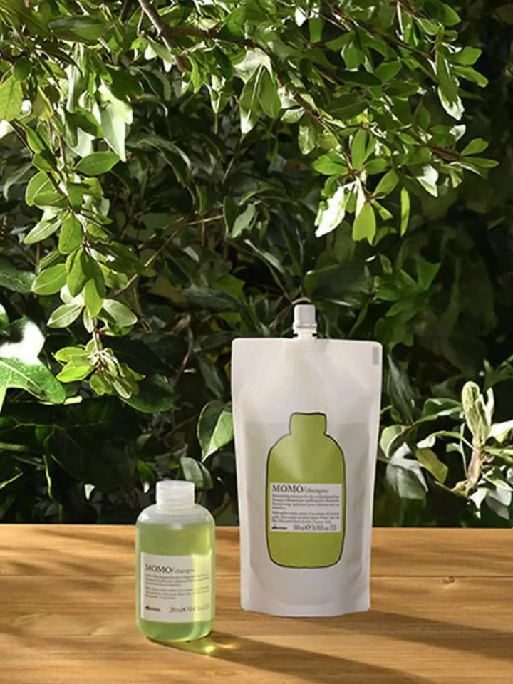 A pouch of Davines MOMO Shampoo Refill and a matching small plastic bottle rest on a wooden table with lush green foliage blurred in the background | Twentyseven Toronto