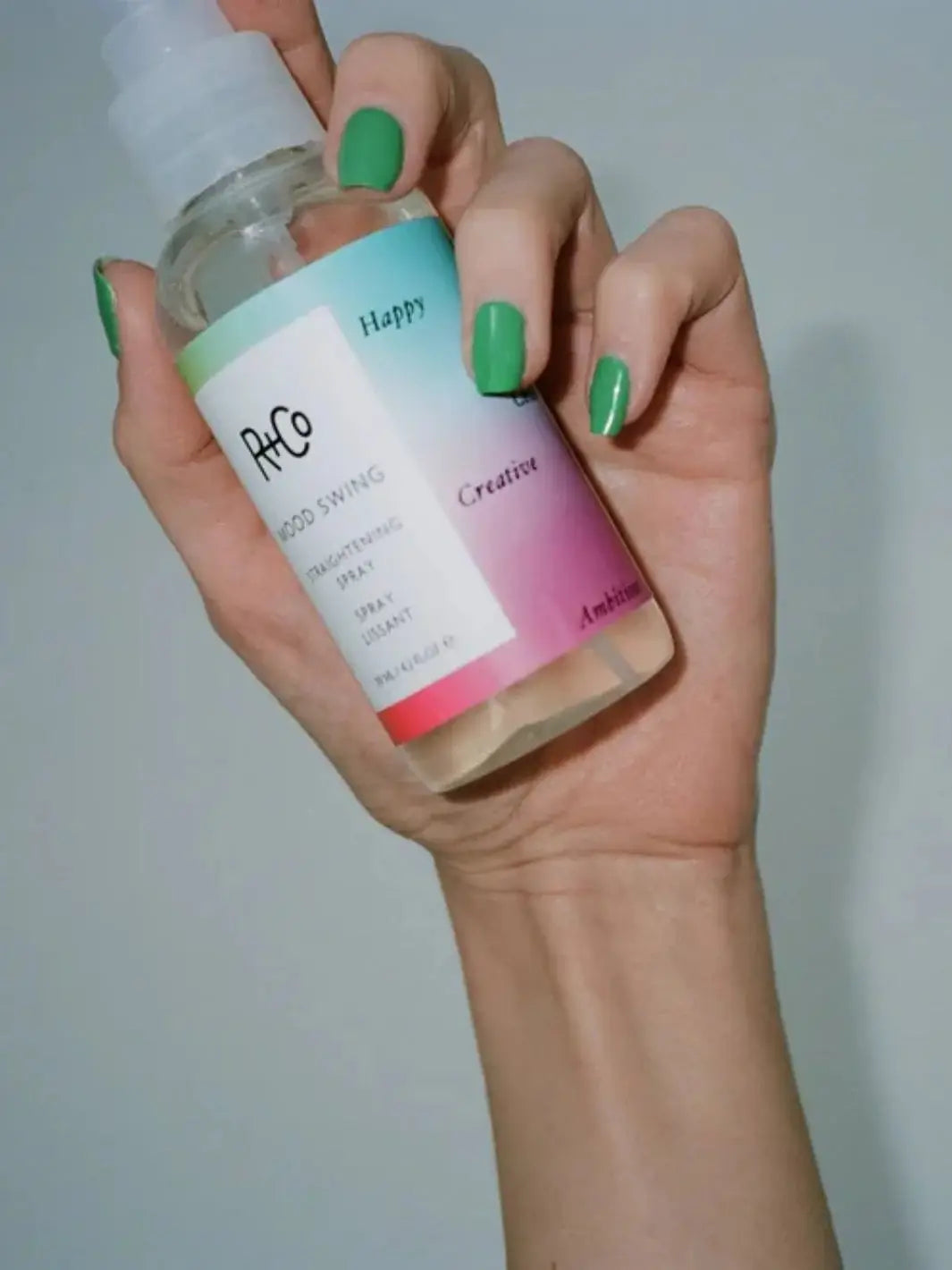 A hand with bright green nail polish holds a clear bottle of R+Co MOOD SWING Straightening Spray against a plain grey background | Twentyseven Toronto