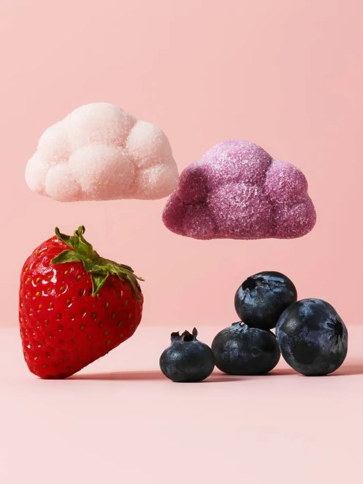One pink and one purple cloud-shaped gummy from the BonBon Swedish Candy Moomin Candy Clouds hover above a fresh strawberry and a small pile of blueberries against a solid pink backdrop | Twentyseven Toronto