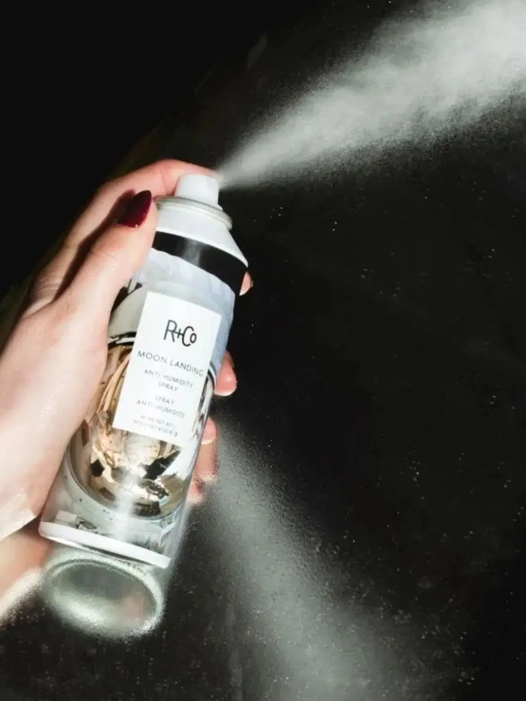 A hand with dark red fingernails presses the nozzle of a can, releasing a stream of mist from the R+Co MOON LANDING Anti-Humidity Spray against a dark background | Twentyseven Toronto
