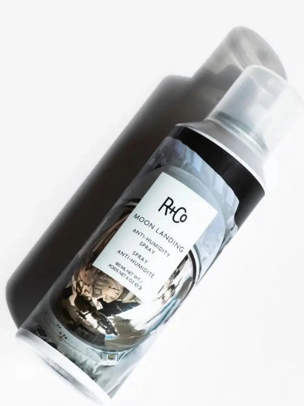 An aerosol bottle of R+Co MOON LANDING Anti-Humidity Spray lies diagonally across a clean white surface, casting a soft shadow | Twentyseven Toronto