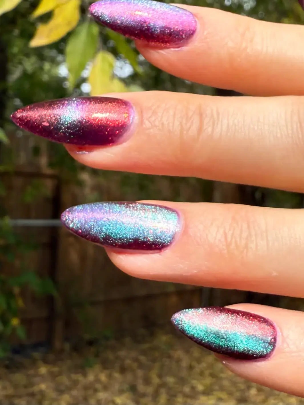 Close-up of a hand with glittery pink and blue 'Mumbling About Apples In Your Dreams' nail polish by Death Valley Nails against a blurred natural background | Twentyseven Toronto