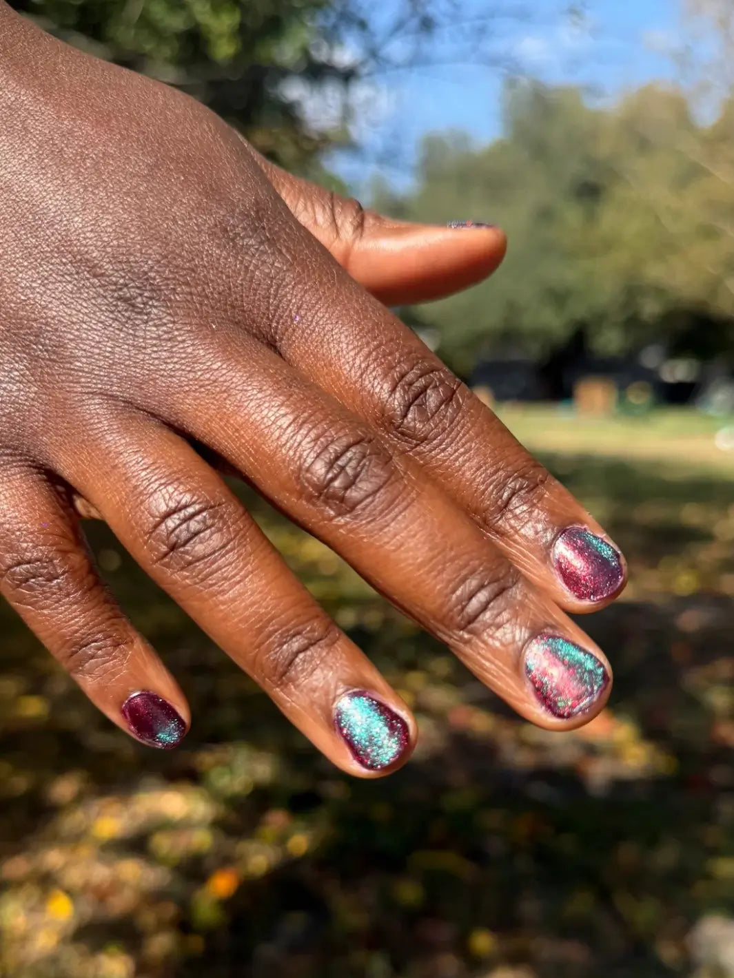 Close-up of a hand with glittery pink and blue 'Mumbling About Apples In Your Dreams' nail polish by Death Valley Nails against a blurred natural background | Twentyseven Toronto
