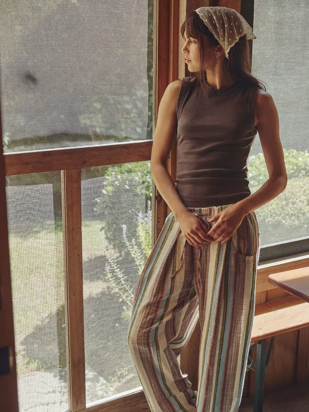 Woman wearing a brown 'Murphy Tank' top in Chocolatier and striped pants from Free People by a window | Twentyseven Toronto