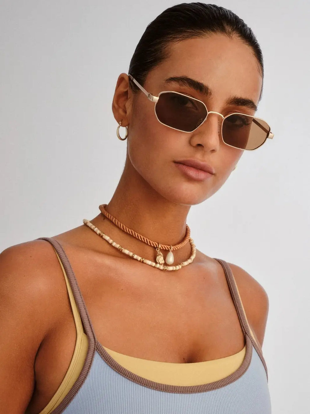 A woman looking forward while wearing the geometric, gold-framed Le Specs Mythic - Gold Light Brown Mono sunglasses paired with layered necklaces | Twentyseven Toronto