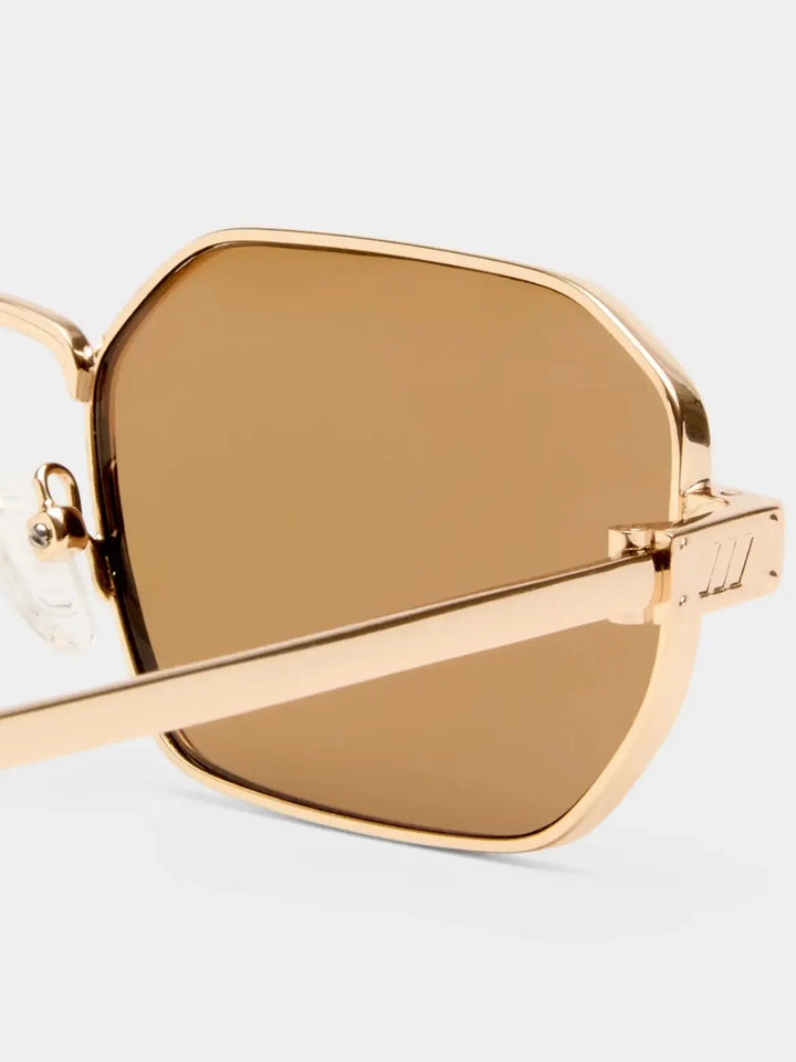 A macro close-up highlighting the angular gold metal frame and light brown lens of the Le Specs Mythic - Gold Light Brown Mono sunglasses at the hinge | Twentyseven Toronto
